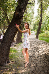 A cheerful young woman in a floral dress stands hugging a large tree in a green, sunlit forest...