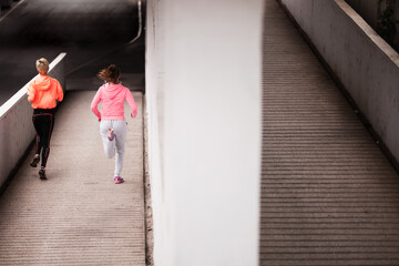Focused on their fitness goals, two women run together on a shaded urban concrete pathway,...
