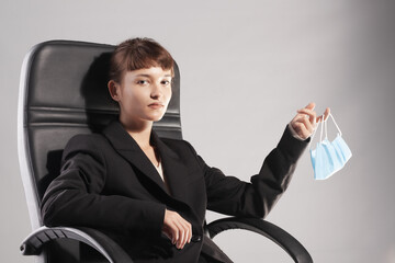 Young businesswoman in office chair holding a medical face mask, contemplating the new normal