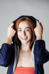Portrait of a cheerful young woman with vibrant red hair adjusting her blue hooded jacket, showcasing a joyful expression.