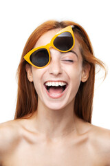 Close-up of a cheerful red-haired young woman wearing yellow sunglasses. Her open, joyous laughter expresses genuine happiness and carefree attitude.