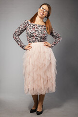 A joyful young woman with glasses wears a floral top and a flowing pink tulle skirt, striking a playful pose on a gray background.