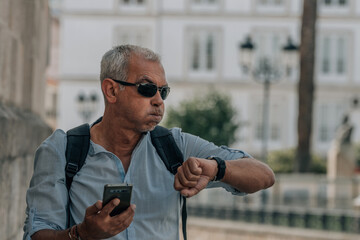 mature man with phone looking with distressed gesture at the clock on the street