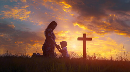Silhouette of mother and child kneeling in prayer together, side view at sunset. Large cross in background. Mother is standing up while the son sits on his knees holding hands with her praying to God