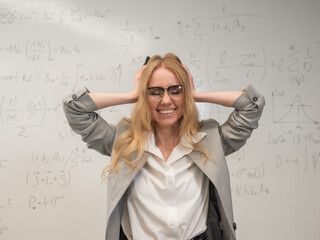 Caucasian woman stands by a white board and holds her head. 