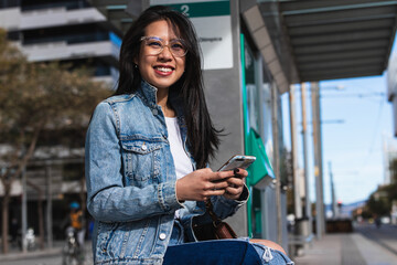 Happy woman uses cell phone while waiting at train station and looking at camera.