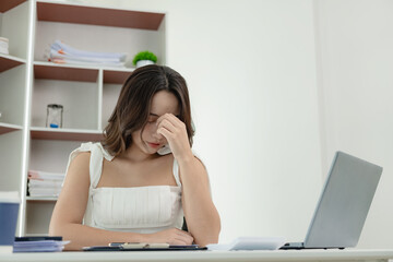 Tired and overworked Asian woman suffers from eye strain, suffers from headaches Holding your head with your eyes closed, thinking about financial mistakes, spending too much.