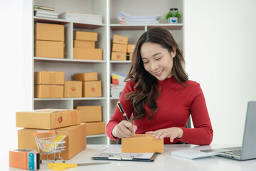 Young Asian business woman smiles and uses laptop to check information on shipping box before sending it to customer. Small business entrepreneur, SME online marketing, delivery concept