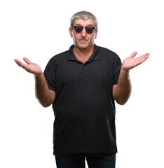 Naklejka premium Handsome senior man wearing sunglasses over isolated background clueless and confused expression with arms and hands raised. Doubt concept.