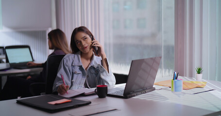 Obraz premium Asian India teen age staff girl sitting desk chair use laptop hold mobile phone talk client help online indoor office Indian busy young adult gen z woman lady use device call centre do job work task