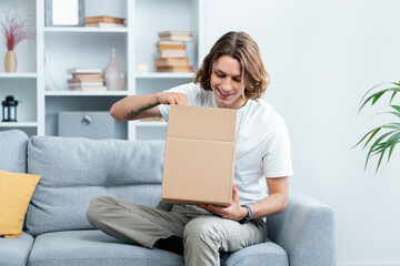 Young Man Smiling While Opening A Cardboard Box On Sofa, Home Delivery Concept Capturing Happiness, Surprise, And Anticipation. Indoor Modern Living Room Setting.
