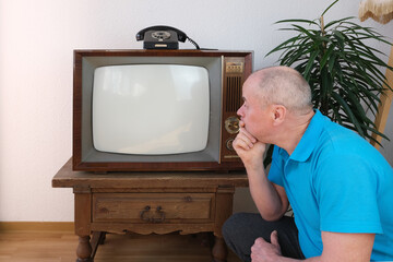 elderly man in blue polo shirt sits on floor in front old retro analog TV, television, white screen...
