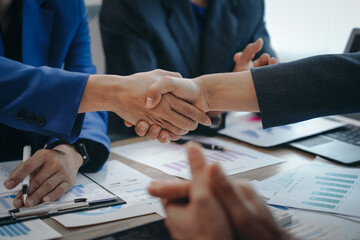 In a business team meeting, success is celebrated with a handshake. Hands close up, dressed in formal suits, working at desks with financial papers, calculators, and laptops.