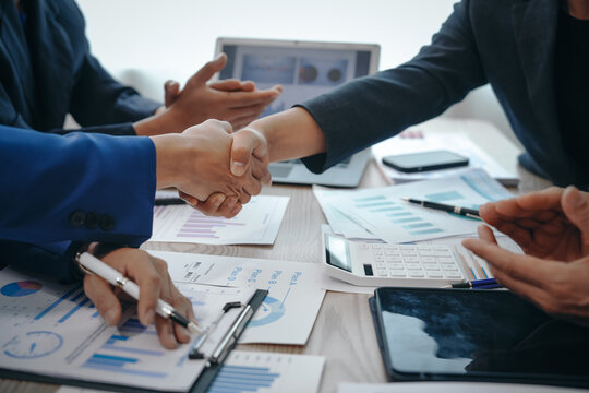 In a business team meeting, success is celebrated with a handshake. Hands close up, dressed in formal suits, working at desks with financial papers, calculators, and laptops.