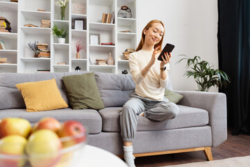 Young Woman Enjoying Time On Sofa At Home, Smiling at Smartphone in Cozy Living Room With...