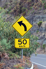 Yellow speed limit and left turn ahead road signs amongst trees and a cliff