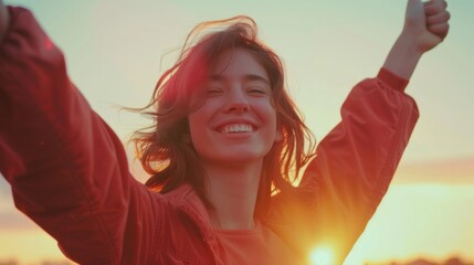 A woman is smiling and holding her arms up in the air. She is wearing a red jacket and is standing in a field