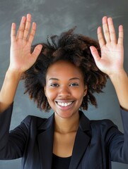 A woman with curly hair is smiling and holding her hands up in the air. She is wearing a black suit and she is happy