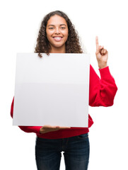 Young hispanic woman holding blank banner surprised with an idea or question pointing finger with...