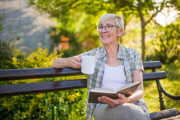 Happy senior woman enjoys reading book and drinking coffee on a bench in her garden.	