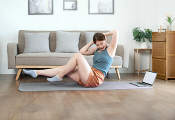 Cheerful young active woman doing fitness at home, using laptop, watching online lessons.