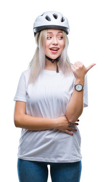 Young blonde woman wearing cyclist security helmet over isolated background smiling with happy face looking and pointing to the side with thumb up.