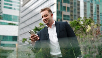 Caucasian business man hold mobile phone and looking at skyscraper view. Skilled male leader standing rooftop in modern building. Caucasian executive manager look at phone while rest his eyes. Urbane.