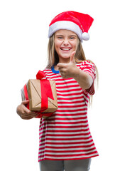 Young beautiful girl wearing christmas hat and holding gift over isolated background happy with big smile doing ok sign, thumb up with fingers, excellent sign