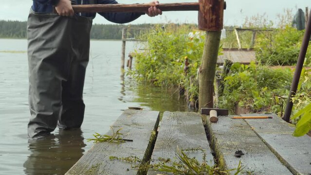 A man in a waterproof suit drives piles into the ground with a giant wooden hammer.