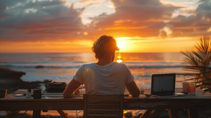 A man is sitting at a table by the ocean, working on his laptop