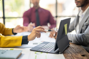A group of people are sitting around a table with a laptop and a pen. One person is pointing at the laptop with the pen. Scene is collaborative and focused