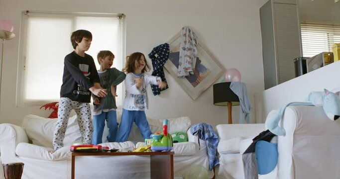 Three kids are energetically jumping on a white couch surrounded by toys and household items in a disordered living room, with sunlight streaming through the window.