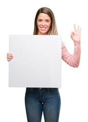 Beautiful young woman holding advertising banner doing ok sign with fingers, excellent symbol