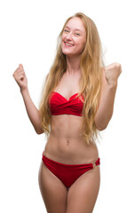 Blonde teenager woman wearing red bikini very happy and excited doing winner gesture with arms raised, smiling and screaming for success. Celebration concept.