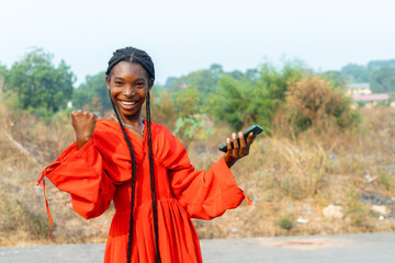 Excited African Woman in Red Dress Holding Mobile Phone, Fist Raised in Joyful Exuberance