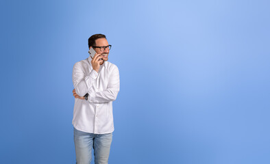 Cheerful male professional in glasses discussing business ideas over mobile phone on blue background