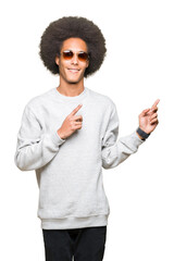 Young african american man with afro hair wearing sunglasses smiling and looking at the camera pointing with two hands and fingers to the side.