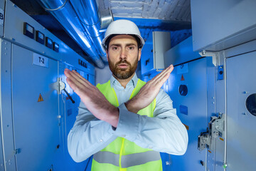Portrait of male electrical engineer technician worker in white hard hat substation worker gestures to halt against a backdrop of blue light, high voltage, and malfunctioning electrical equipment.