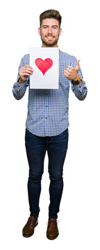 Young handsome man holding paper with red heart happy with big smile doing ok sign, thumb up with fingers, excellent sign