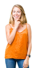 Beautiful young woman wearing orange shirt over isolated background looking confident at the camera with smile with crossed arms and hand raised on chin. Thinking positive.