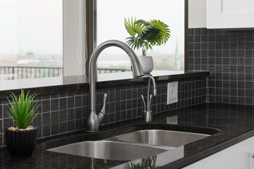 Kitchen sink detail shot with a square dark grey tile backsplash, black granite countertop, white...
