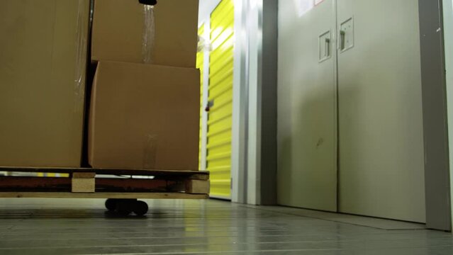 A mechanical loader with cardboard boxes pulls up to the doors of freight elevator in the self-storage warehouse. Close-up view from low angle. 4K