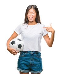 Young asian woman holding football soccer ball over isolated background with surprise face pointing finger to himself