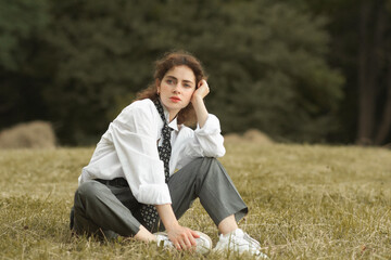 A girl in a white shirt and tie sits pensive in the field. Grain effect used