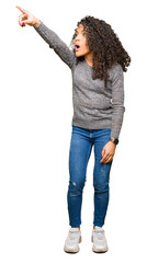 Young beautiful woman with curly hair wearing grey sweater Pointing with finger surprised ahead, open mouth amazed expression, something in front