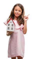 Obraz premium Brunette hispanic girl holding little house very happy pointing with hand and finger to the side