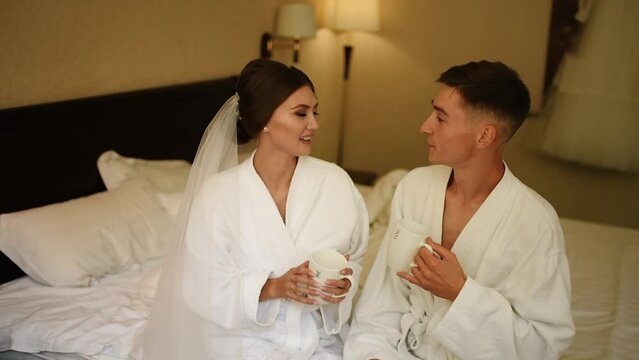 Bride and groom barefoot drinking coffee cups in bed at morning together. Couple hugging smile while lying bed. Woman in veil and white bathrobe with man with bare feet having fun and rejoicing kiss