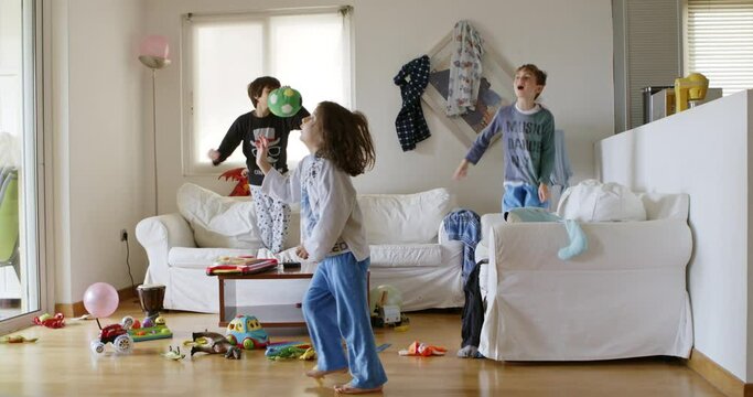 Three kids are energetically jumping on a white couch surrounded by toys and household items in a disordered living room, with sunlight streaming through the window.
