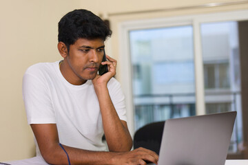 young indian man using laptop and talking on the phone at home
