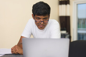 indian male student studying at home using laptop computer and writing notes